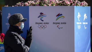 A woman wearing a face mask to protect against Covid-19 sits near landscaping decorated with the logos for the Beijing Winter Olympics and Paralympics on a pedestrian shopping street in Beijing