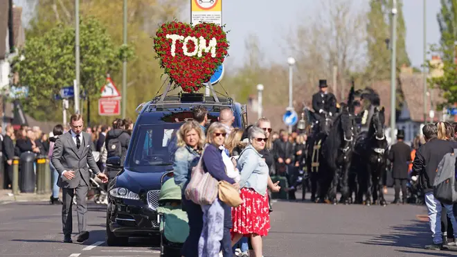 A heart-shaped red rose piece spelling "Tom" adorns the hearse