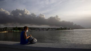 A resident sits on the sea wall as smoke rises in the background from a deadly fire at a large oil storage facility in Matanzas, Cuba