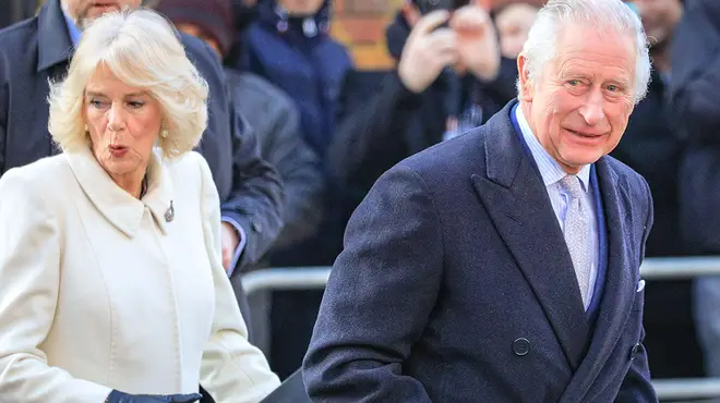 King Charles and wife Camilla on a royal event where she wears a cream coat and black leather coat and Charles wears a blue coat and shirt
