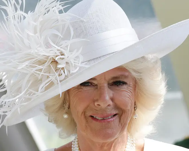 Queen Camilla smiling while wearing a white hat with a feather feature