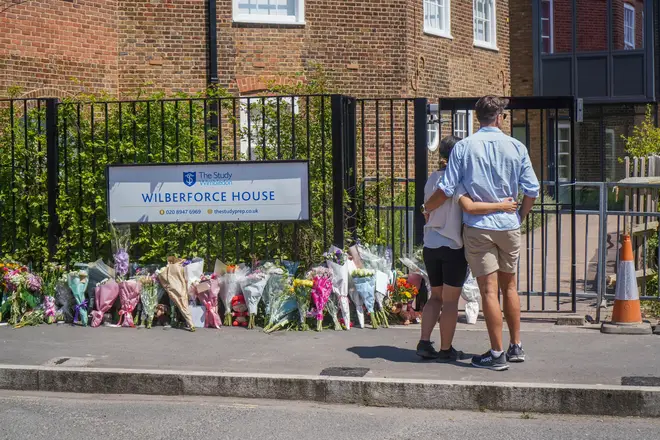A couple stand next to the floral tributes at the gates of the school