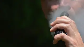 A man vaping with focus on his hand as he holds the e cigarette / vape mod up to his mouth while exhaling vapour